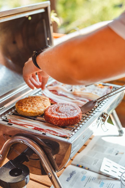 Person cooking burgers and bacon on an outdoor flat top griddle in a backyard setting